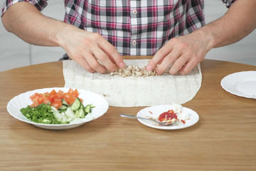Man is cooking shawarma on the kitchen table at home. Pita, vegetables and green onion with sauce and mayonnaise. Close-up hands.