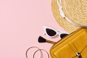 Top view of straw hat, bracelets, cosmetic brush, sunglasses and yellow bag on pink background
