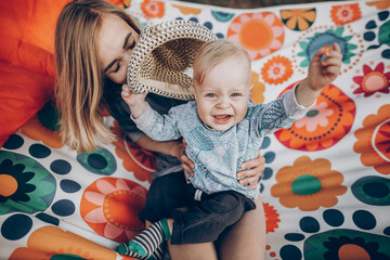 Young family portrait of a beautiful hipster mother with cute blonde boy with blue eyes and vintage hat sitting on a boho hammock outdoors, family camping vacation concept