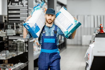 Portrait of a handsome workman in blue overalls holding bags with construction mixture in the building supermarket