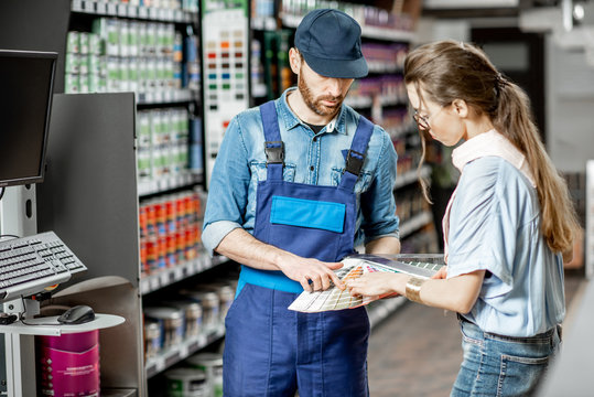 Young woman client choosing paint from color swatches, standing with workman in supermarket with building goods