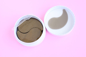 Obraz na płótnie Canvas Round white container with black golden anti aging collagen lifting patches under eyes for woman face care with selective focus on pink neutral background. Hydrating beauty facial anti wrinkles patch