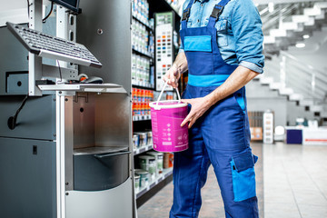 Workman in uniform putting bucket with paint into the machine for coloring in the building shop