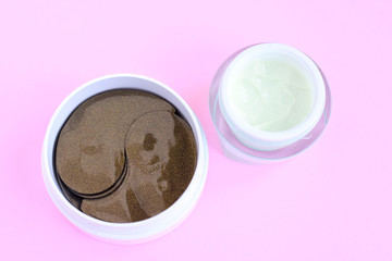Plastic beauty jar with green face cream and container with black golden anti aging collagen lifting patches under eyes for woman face care with selective focus on pink neutral background. 