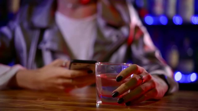 woman is typing message on smartphone touch screen and holding glass with vodka in nightclub
