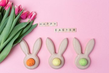 Easter holiday background with colored eggs in bunny ears shaped egg cups, pink flowers tulip , Happy Easter wooden letters. Flat lay.