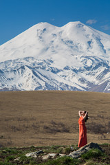 brunette woman stand in long red dress on the background of high snow-capped mountains 