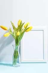 A bouquet of yellow tulips in a glass vase and wooden white frame on the table.