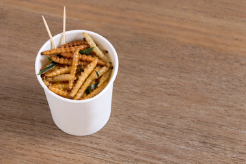 Bamboo Caterpillar insects for eating as food. Worm deep-fried crispy snack in disposable cup for take-away home on wood table background. It is good source of protein edible. Entomophagy concept.