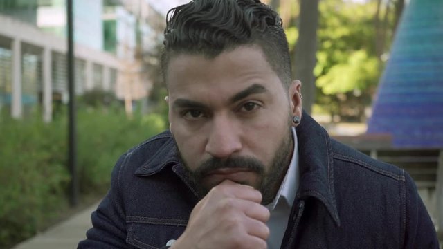 Serious bearded man in jacket looking at camera. Portrait of handsome man with beard and piercing sitting with hand on chin outdoor. Facial expression concept