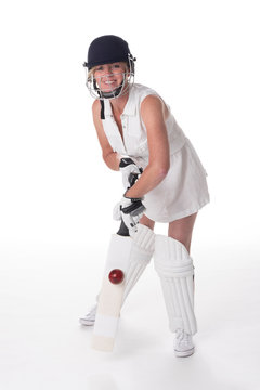 Woman cricketer in a white dress with a safety helmet, shin pads, a bat and ball.