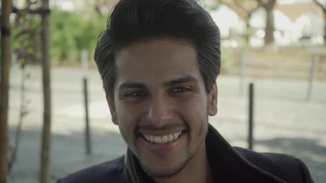 Portrait of handsome young man smiling at camera. Head and shoulders view of cheerful Indian man laughing and looking at camera on street. Emotion concept