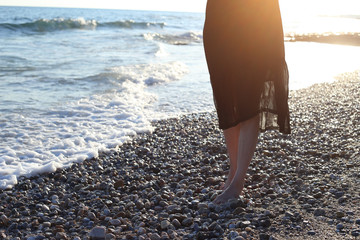 Young girl in black dress walking alone on the sandy tropical beach at sunset. Close Up detail of female feet and golden sand on beach.