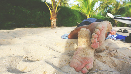 Close up of crossed sandy feet of a man relaxing and sleeping on a beautiful beach on the tropical island, with palm trees in the background