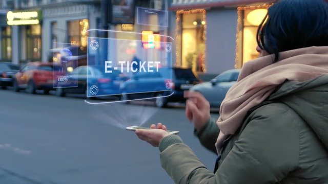 Unrecognizable woman standing on the street interacts HUD hologram with text E-ticket. Girl in warm clothes with a scarf uses technology of the future mobile screen on background of night city