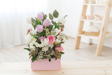 Spring flower bouquet with tulips, roses, freesia and eucalyptus leaves in pink wooden box stands on wooden table in the living room.