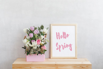 Frame with text HELLO SPRING and spring flower bouquet with tulips, roses, freesia and eucalyptus leaves in pink wooden box on a wooden table on a background of light gray walls. Home interior decor.