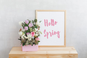 Frame with text HELLO SPRING and spring flower bouquet with tulips, roses, freesia and eucalyptus leaves in pink wooden box on a wooden table on a background of light gray walls. Home interior decor.