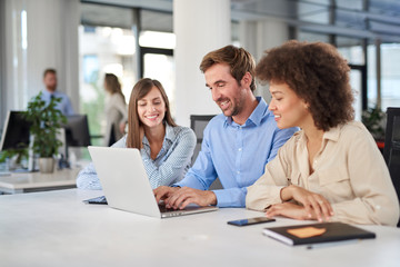 Coworkers sitting at table and solving problem. Man using laptop while two female colleagues looking at laptop and helping him..