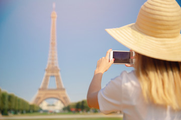 Fototapeta premium Travel blogger young woman in hat takes photograph of Eiffel Tower on telephone in Paris, France