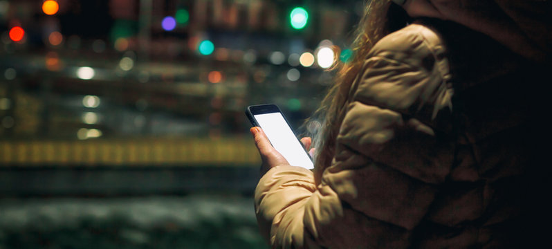 The young woman holds her phone and read some text in the evening city in winter