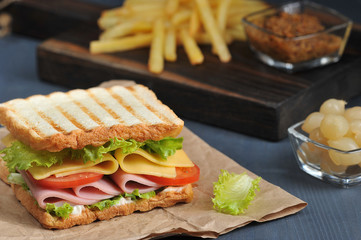 A ham and cheese sandwich takes up the entire frame space. In the background are french fries, pickled onions and mustard. Dark background. Close-up. Macro shooting.