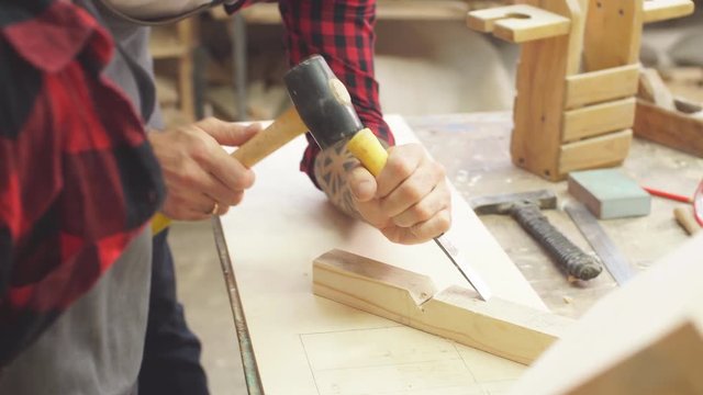 Man in apron works with chisel in a carpentry shop. Carpenter holding a chisel in wooden workshop