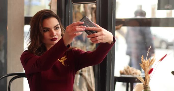 Gorgeous young brunette model with bright make up and lips suits the color of her dress making selfies on her smartphone sitting in cafe at lunch.