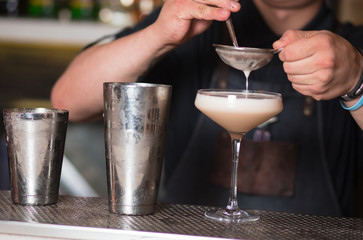 Cocktail being poured at a bar preparing a drink. Motion blur of the bartender's hand and strainer. Selective focus on the foreground glass. Blurred people in the background.  Trendy black stylish 