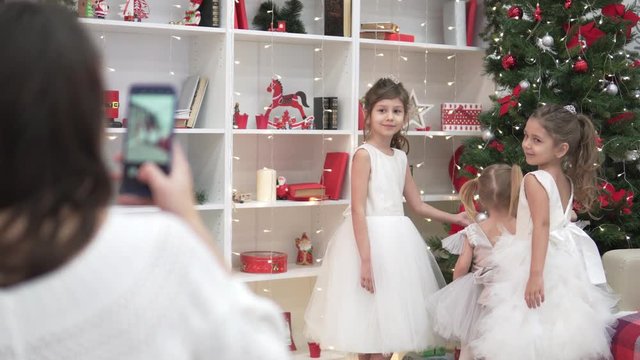 Mom takes pictures of smart daughters at the Christmas tree. Big family is preparing to celebrate Christmas. Fancy girls are standing by the Christmas tree, a woman is making a photo of children on th