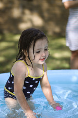 A little girl plays with colored balls in an inflatable pool, while her mother looks at her thoughtfully. The lawn, in the garden, at home. Splashes of water, refreshment from the summer heat.