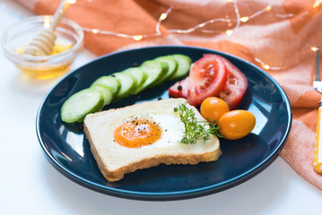 Homemade egg in a hole with toast bread black peppers on it and vegetables on a blue ceramic plate with honey on a white background, close up