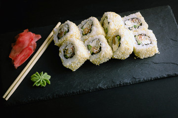 Sushi rolls, wasabi, pickled ginger and chopsticks on a black plate.