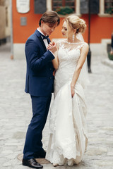 Naklejka premium luxury wedding couple walking at old building on square in light. stylish bride and groom embracing in city street. romantic sensual moment.