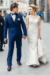 stylish bride and groom walking in city street. happy luxury wedding couple holding hands in light . romantic sensual moment.   man and woman looking at each other