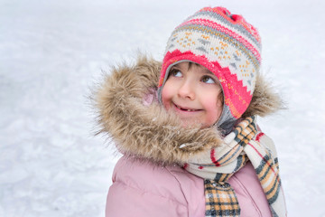 A portrait of a little toothless girl in a purple down puffy jacket and a hat in winter. Looking upwards.