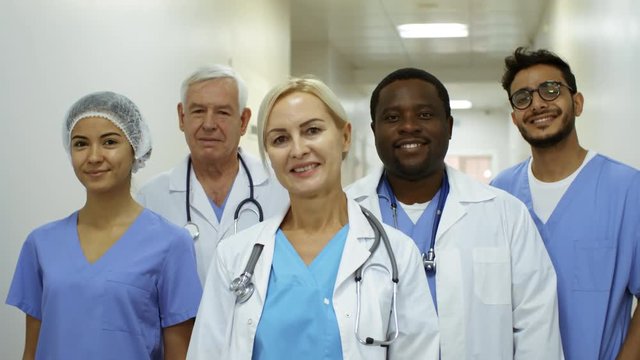 Dolly shot of team of medical professionals wearing scrubs and lab coats standing in hospital corridor and smiling for camera