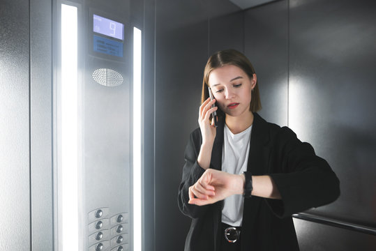 Young european businesswoman is talking on her phone and checking time on her watch in the elevator. Employee using her smartphone and watching her clock in the lift being in hurry.