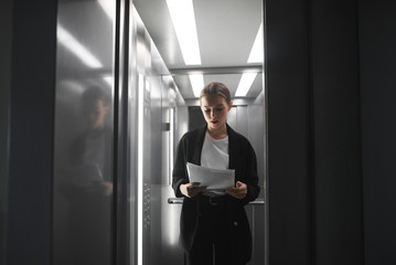 Busy concentrated office worker is reading the documents while the elevator door is closing. Young businesswoman is checking the documentation is the lift when the door is closed.
