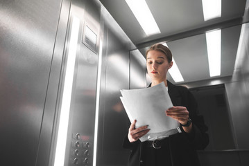 Curious corporate businesswoman standing in the elevator skeptically looking at the documents....