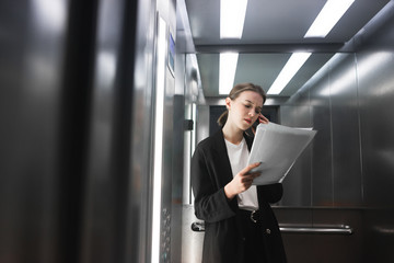 Serious female office worker talking on her smartphone and checking the documentation in the elevator. Young businesswoman reading the papers and communicate by phone.