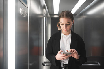 Thoughtful young female office worker is using her smartphone and listening to music in headphones in the lift. Portrait of concentrated businesswoman texting on her phone in the elevator.