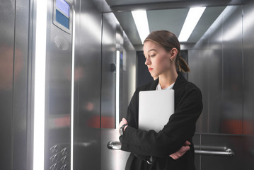 Young businesswoman is concentrated on lookinig at the floor number in the elevator holding her laptop. Female office worker in the lift is keeping the notebook in her hands looking aside. © bodnarphoto