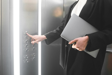 Female employee pressing the elevator button and holding the laptop. Businesswoman in the formal black suit is pushing the button of the lift and keeping her notebook. © bodnarphoto