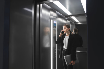 Young businesswoman is talking on the phone in the elevator holding the laptop in her hand. Female...