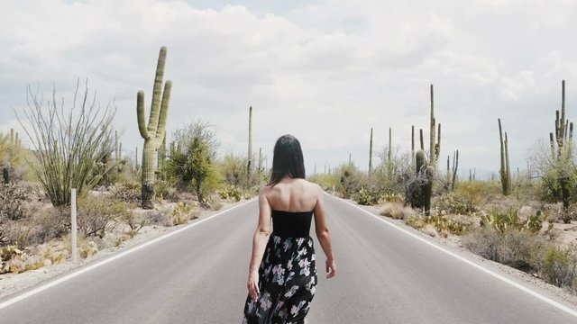 Slow motion camera follows young happy tourist woman lost walking along hot desert road in Saguaro cactus national park.