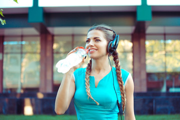 Modern young sportswoman wearing headphones about to drink water from bottle on street