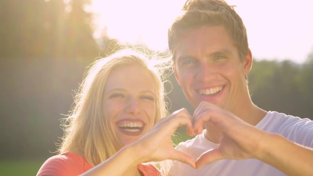 SLOW MOTION, PORTRAIT, LENS FLARE: Young woman laughs while making a heart shape with her boyfriend. Adorable couple enjoying their date in the sunny nature and makes a heart symbol with their fingers