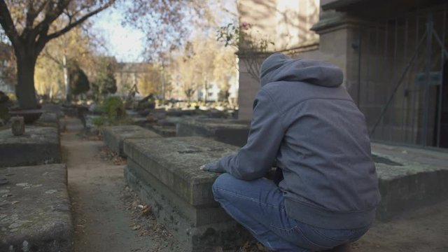Sad man praying near grave on ancient cemetery, commemorating family, generation