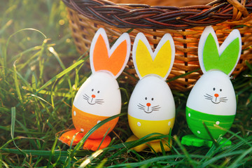Handmade funny creative eggs lies on green grass next to natural basket at sunny spring day. Happy Easter! Egg hunt, decoration for Easter.